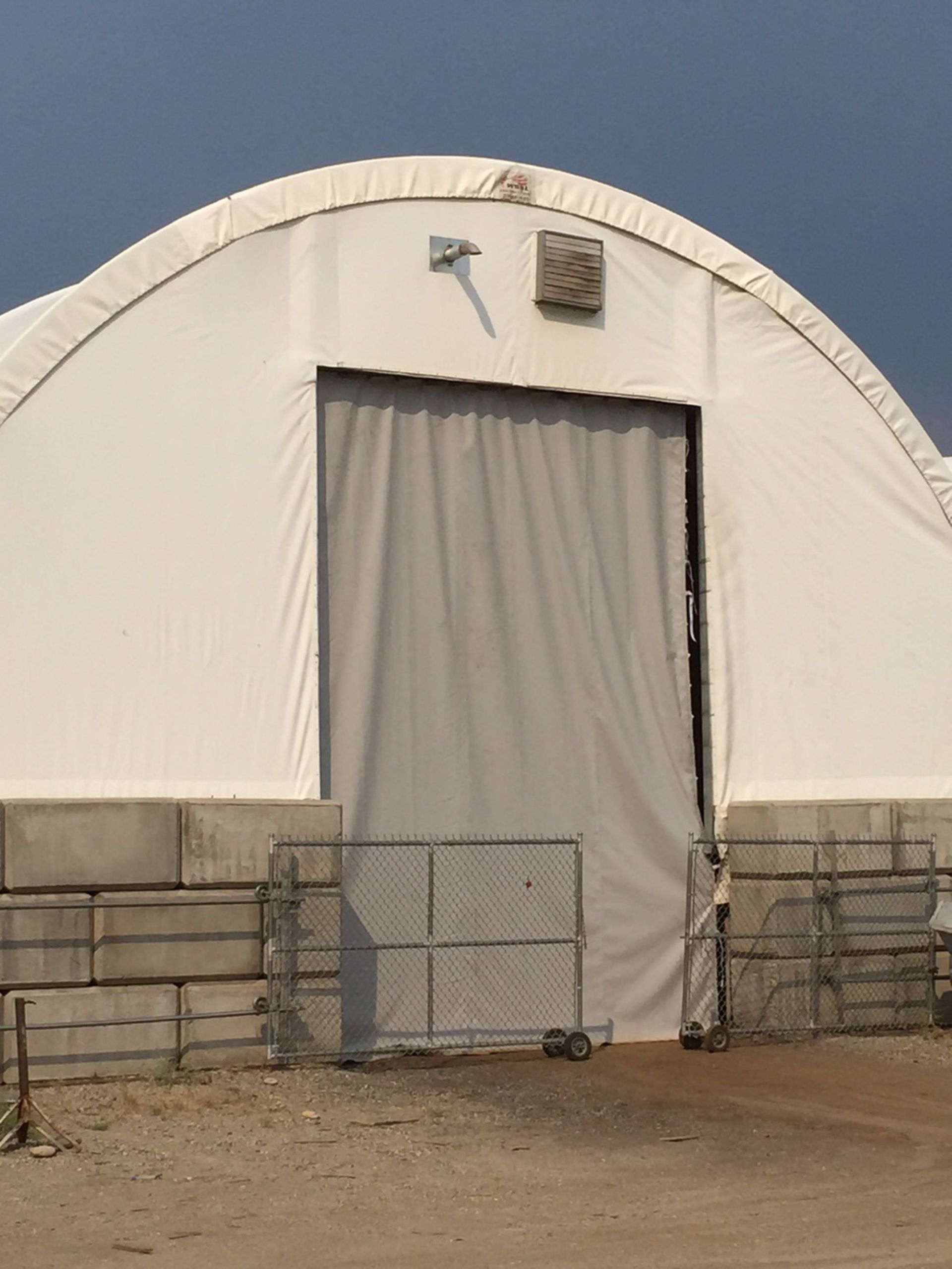 White arched-roof building with a large gray curtain door, a gray metal gate, and concrete blocks along the base.