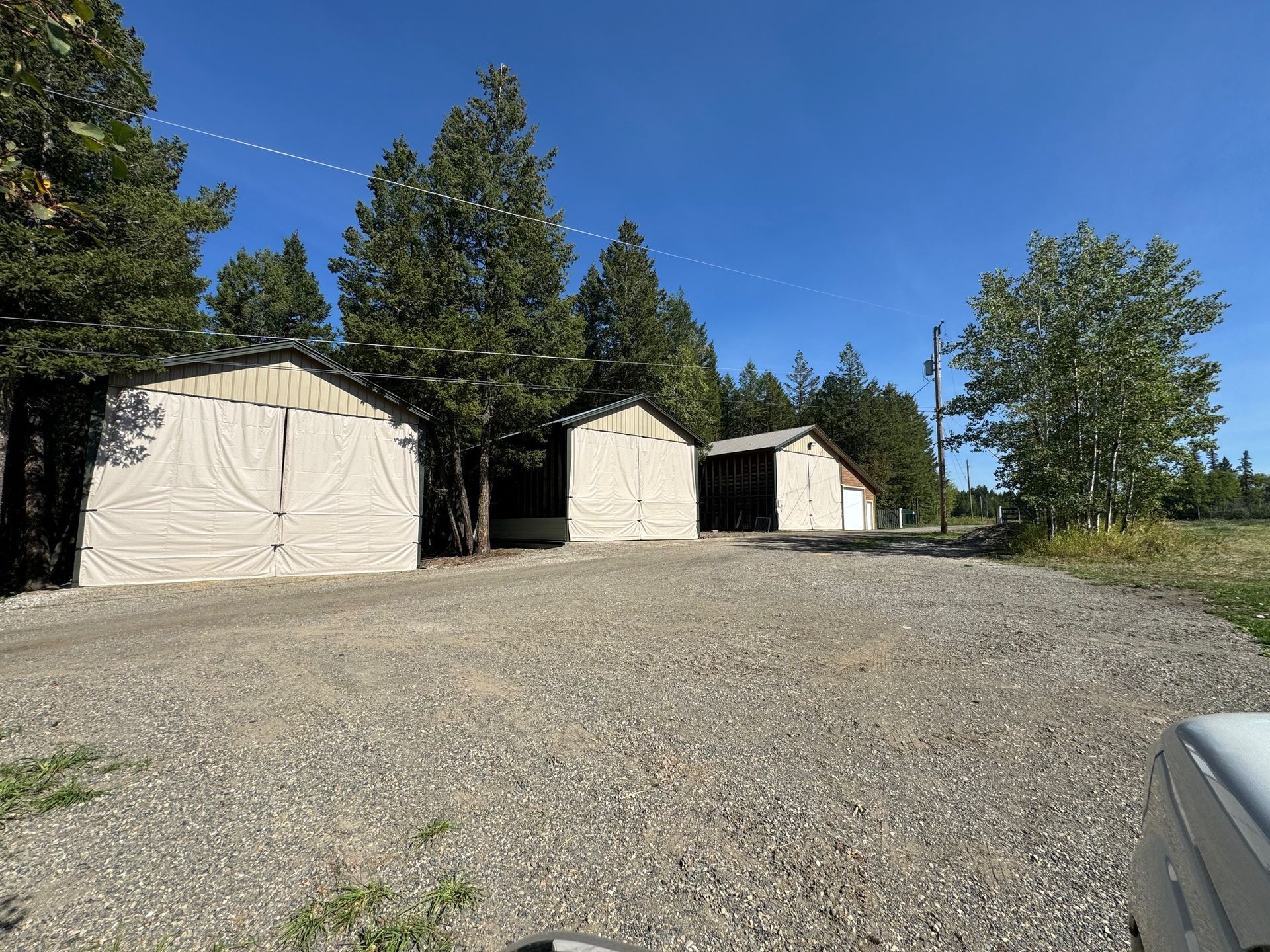 Three tan storage sheds with open doorways on a gravel driveway, beneath a blue sky.
