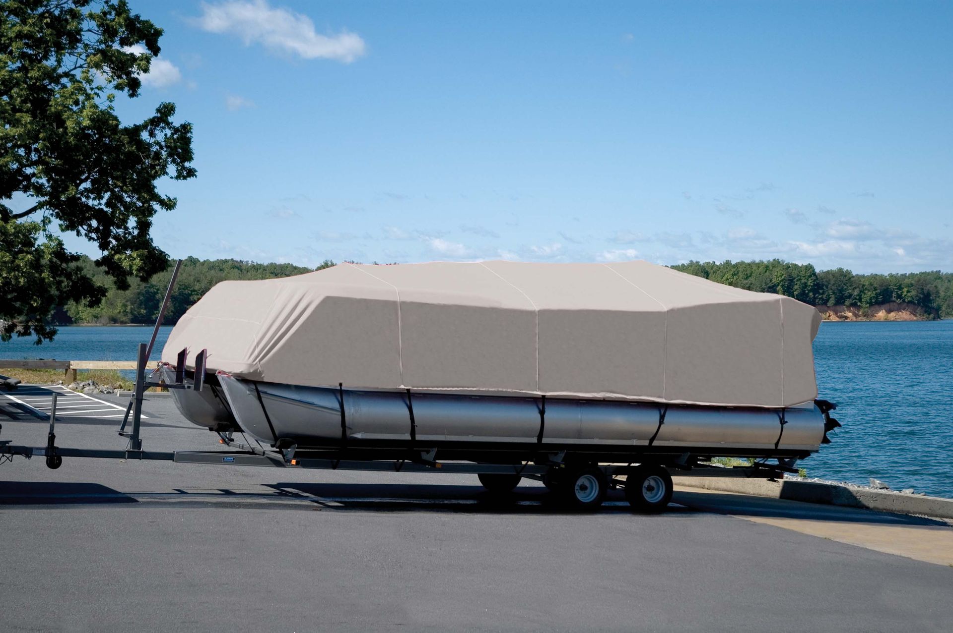 Pontoon boat on a trailer, covered with a beige tarp, parked near a lake.