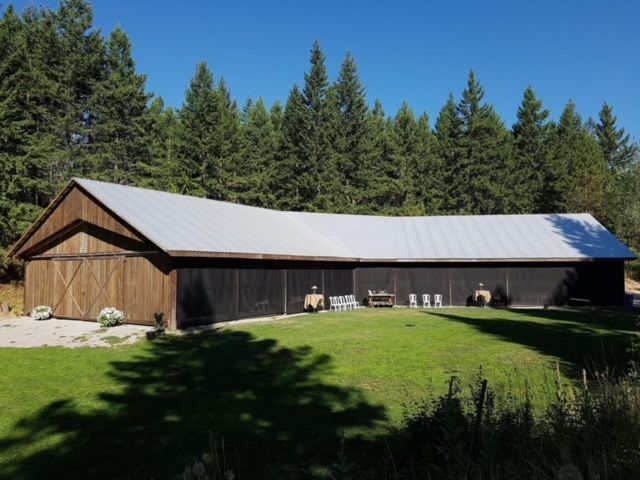 Wooden barn with a silver roof on a grassy field with trees under a blue sky.