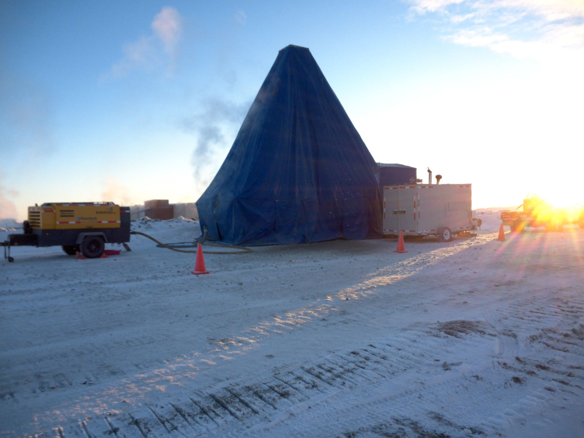 A structure covered in blue tarp with equipment on a snowy, sunny landscape.