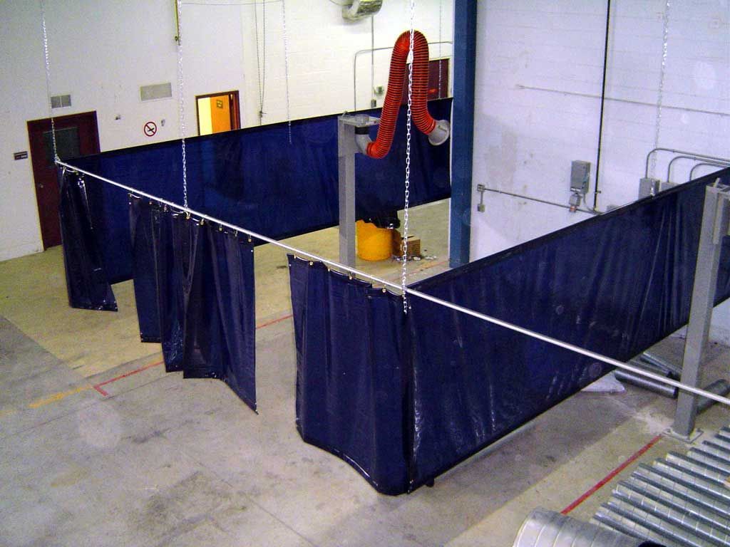 Blue welding curtains enclose a work area in a factory, with a red exhaust hose visible.