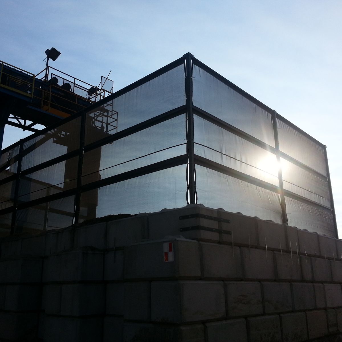 A metal mesh enclosure sits atop concrete blocks, with a conveyor belt in the background and bright sunlight.