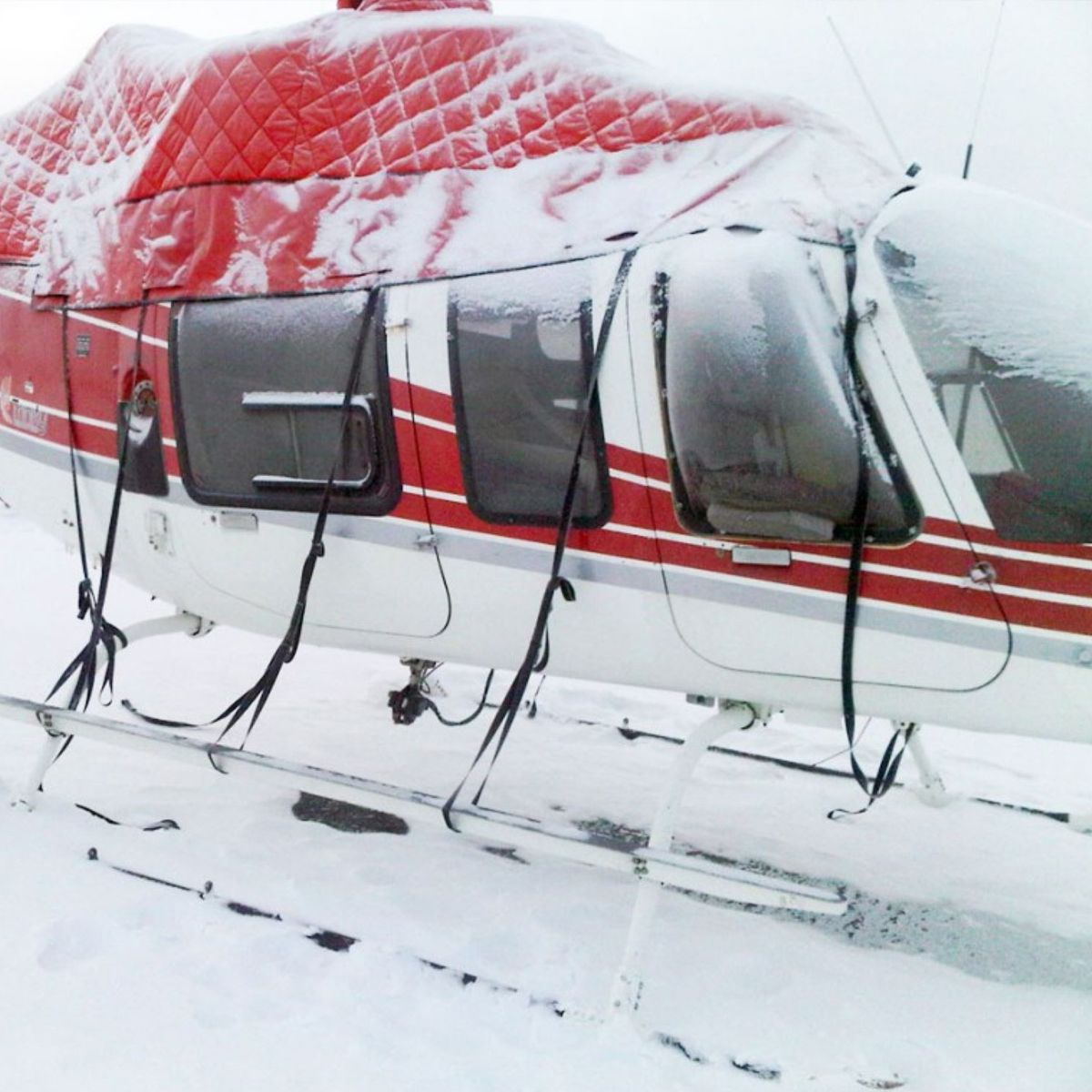 Snow-covered red and white helicopter with window covers secured by black straps, in a snowy setting.