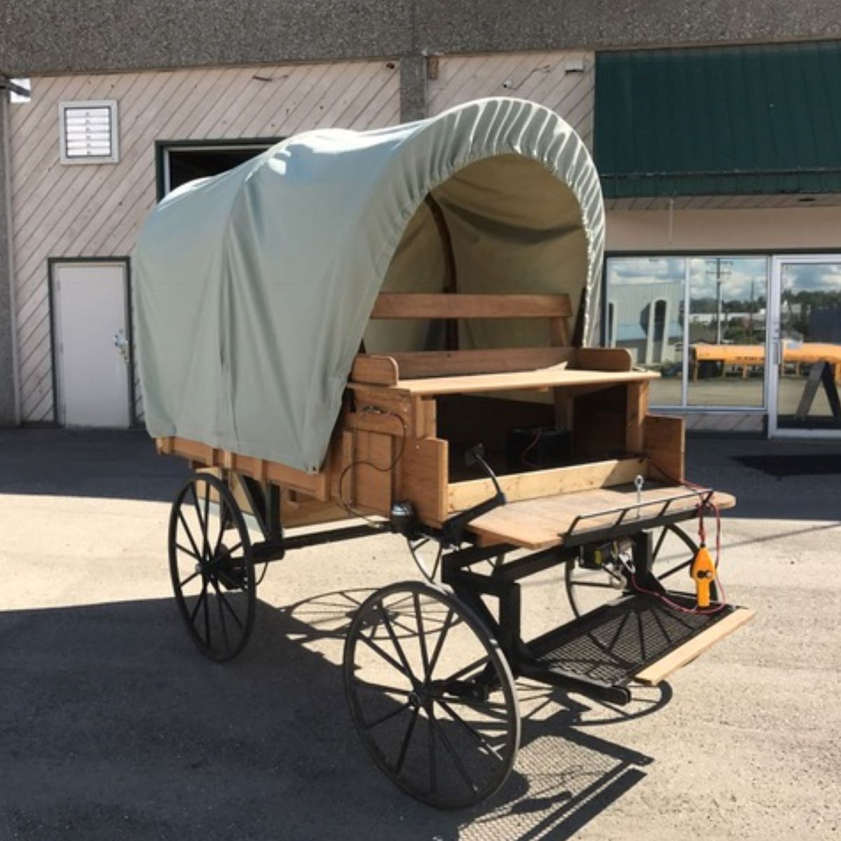 Covered wagon on a paved surface in front of a building. Light green canvas cover, wooden construction, black wheels.