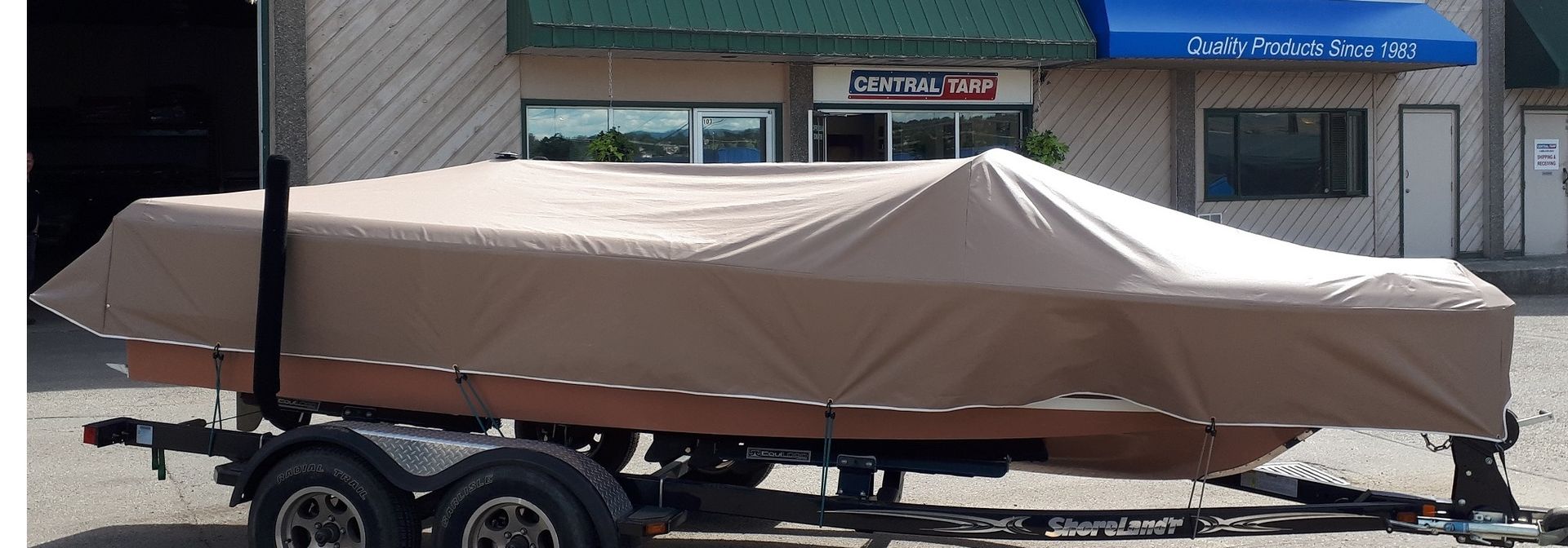 Boat on a trailer, covered with a tan tarp, parked in front of a building.