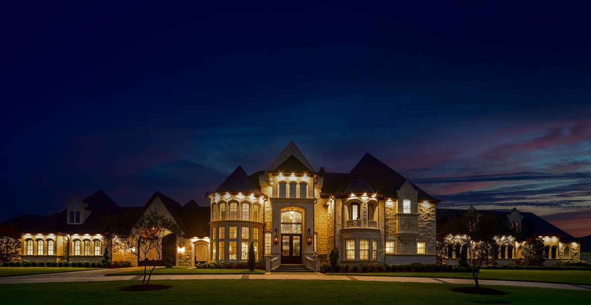 Large, illuminated stone mansion at dusk with manicured lawn.