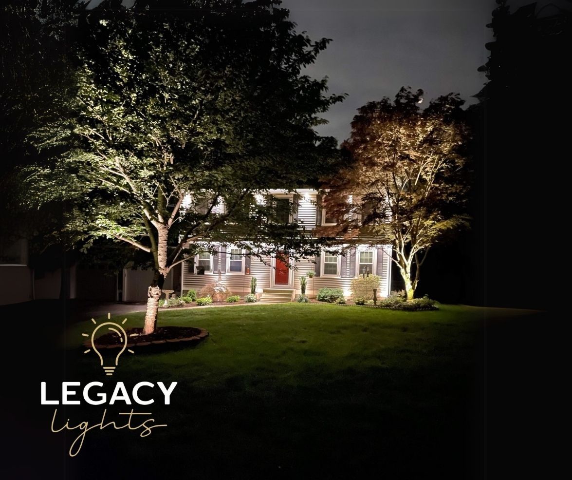 A house at night, illuminated by landscape lights. The lawn is green, trees are lit, and the sky is dark.