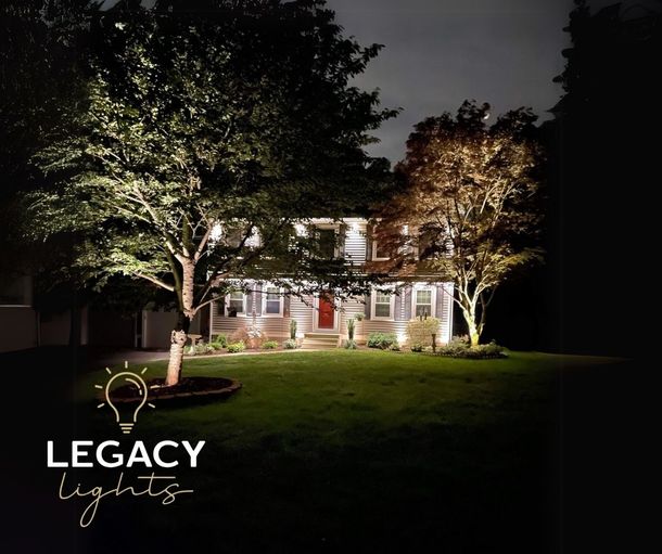 A house at night, illuminated by landscape lights. The lawn is green, trees are lit, and the sky is dark.