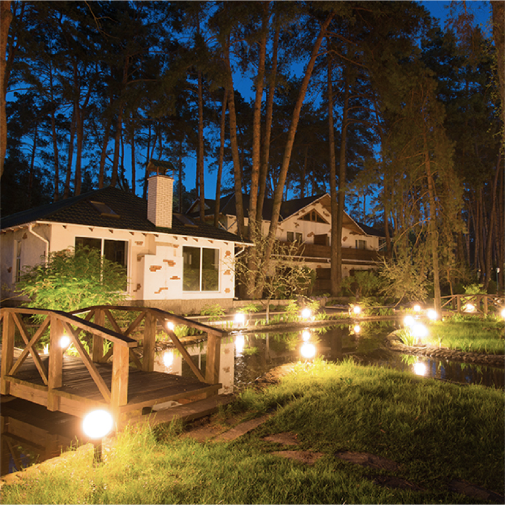 Wooden bridge and glowing lights lead to houses nestled in a dark forest at night.