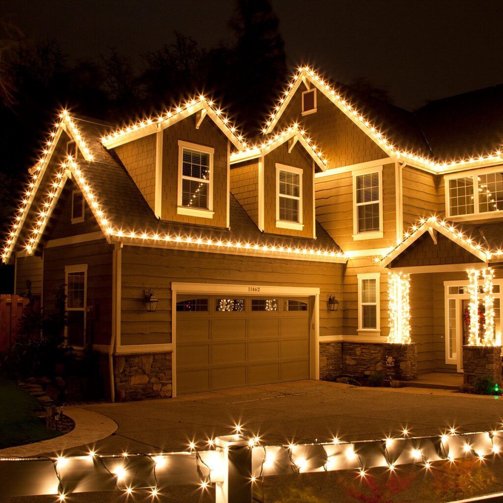 House decorated with warm white Christmas lights; lit roofline, fence, and pillars.
