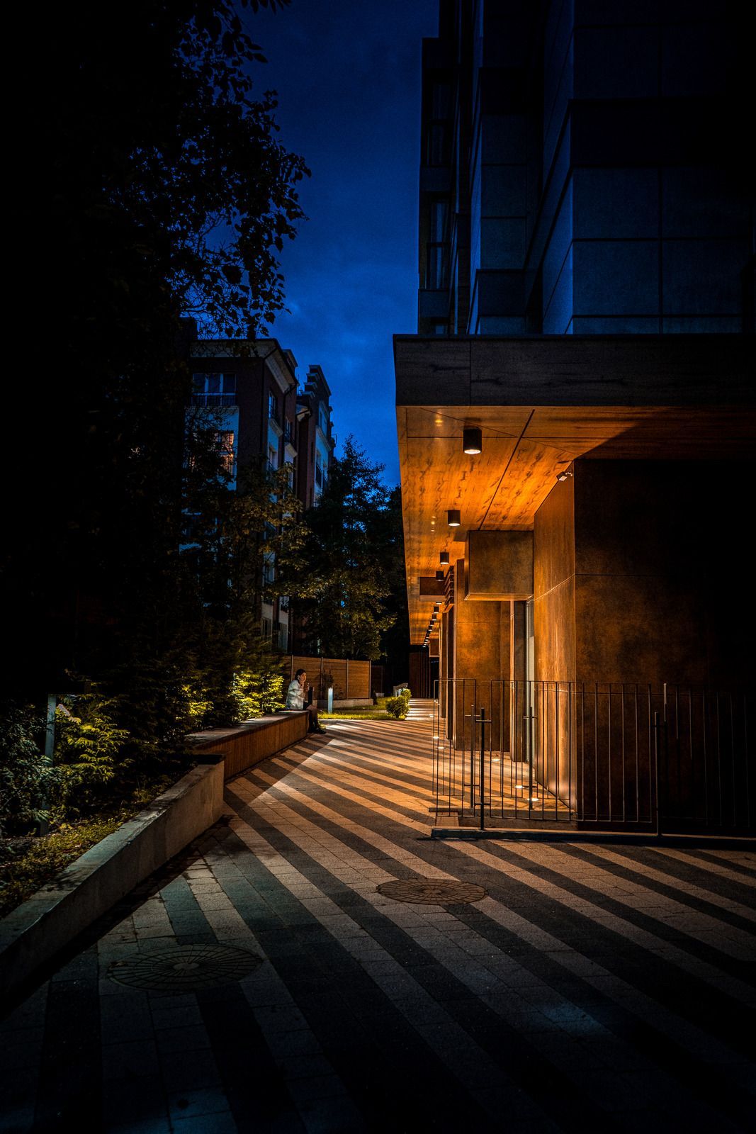 Nighttime view of a modern building entrance with shadow stripes on walkway. Dark blue sky with warm building lighting.