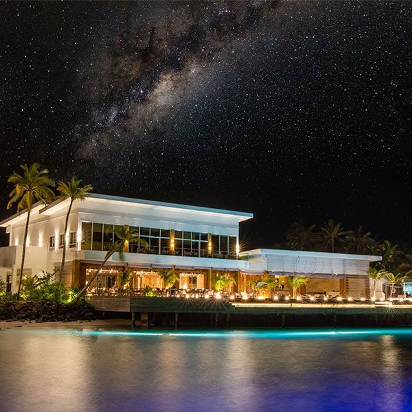 Restaurant at night under the starry Milky Way, with ocean and palm trees.
