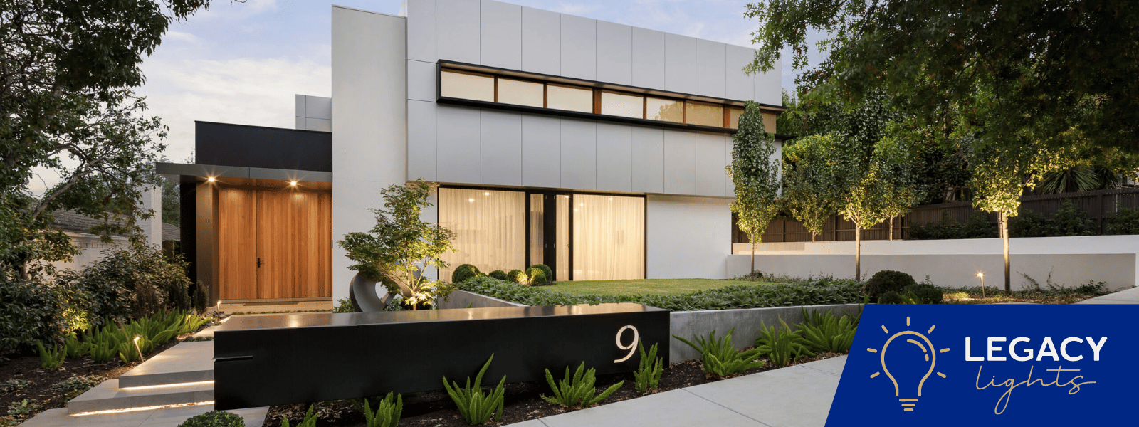 Modern house with exterior lighting. Concrete walkway leads to wooden door. Lawn with small trees.