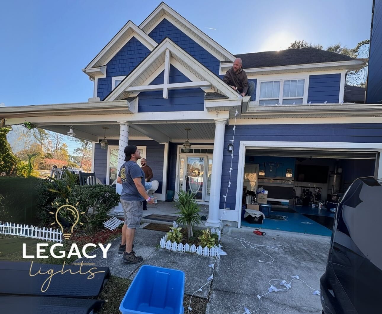 Workers install lights on a blue house with a white trim; one on the roof, two on the ground, with a blue bin.