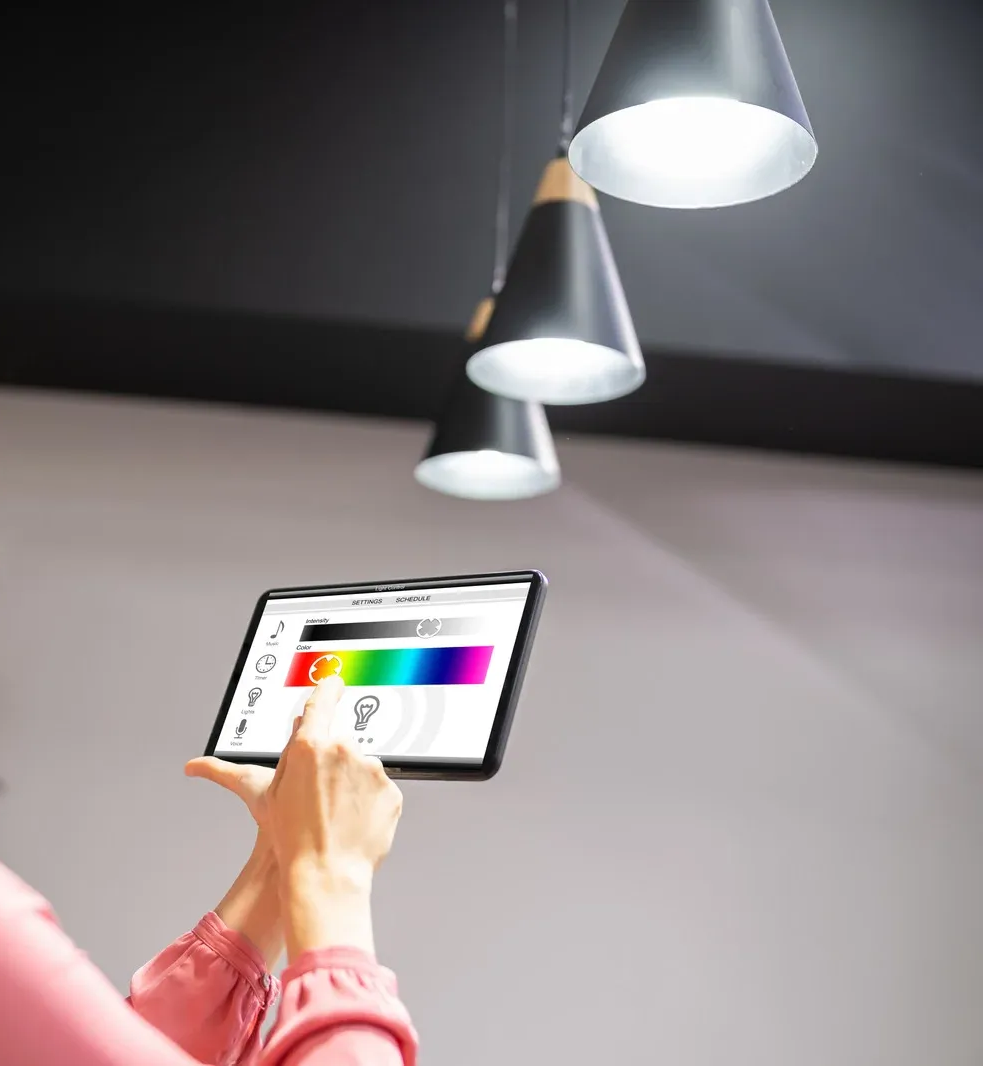 Person using a tablet to control smart lights hanging from a black ceiling, adjusting colors.