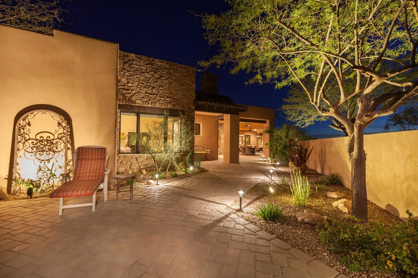 Night exterior of a house with a decorative arched door, paved walkway, and lit landscaping.