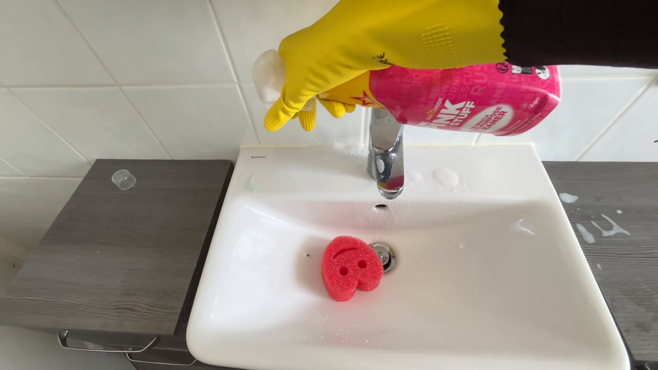 Gloved hand holding pink cleaning spray above a white bathroom sink with a red drain stopper