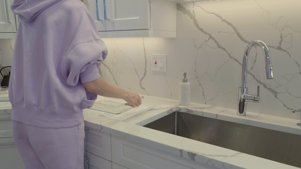 Person in lavender top at a kitchen sink, washing hands beside a stainless-steel basin.