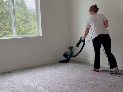 Person vacuuming a carpeted room near a window with a blue vacuum cleaner.