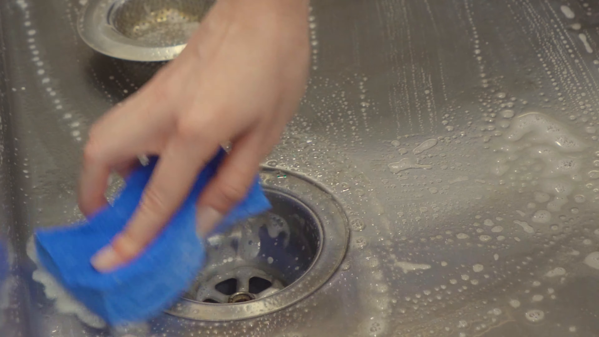 Hand cleaning a stainless steel sink with a blue sponge and soapy water