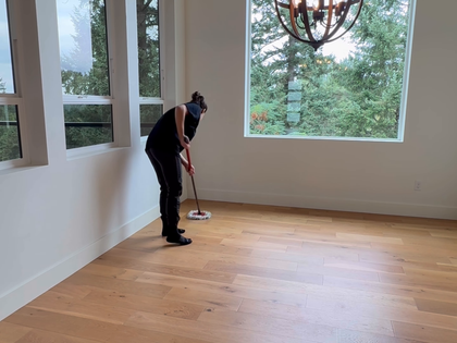 Person mopping a wooden floor in a bright room with large windows and a chandelier.