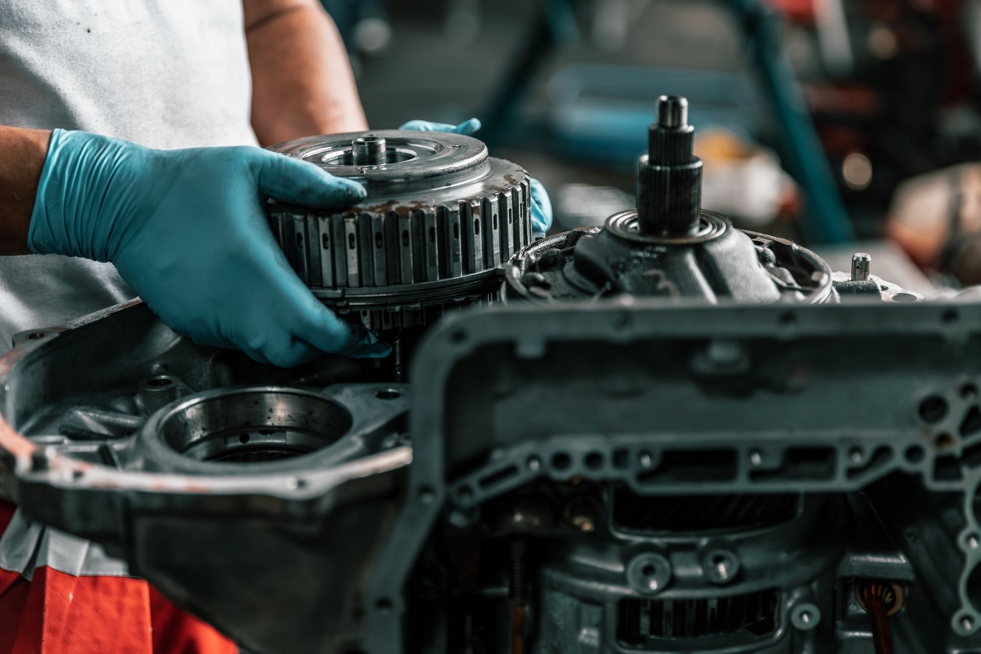Gloved hands working on a car engine with metal parts and gears visible in a garage