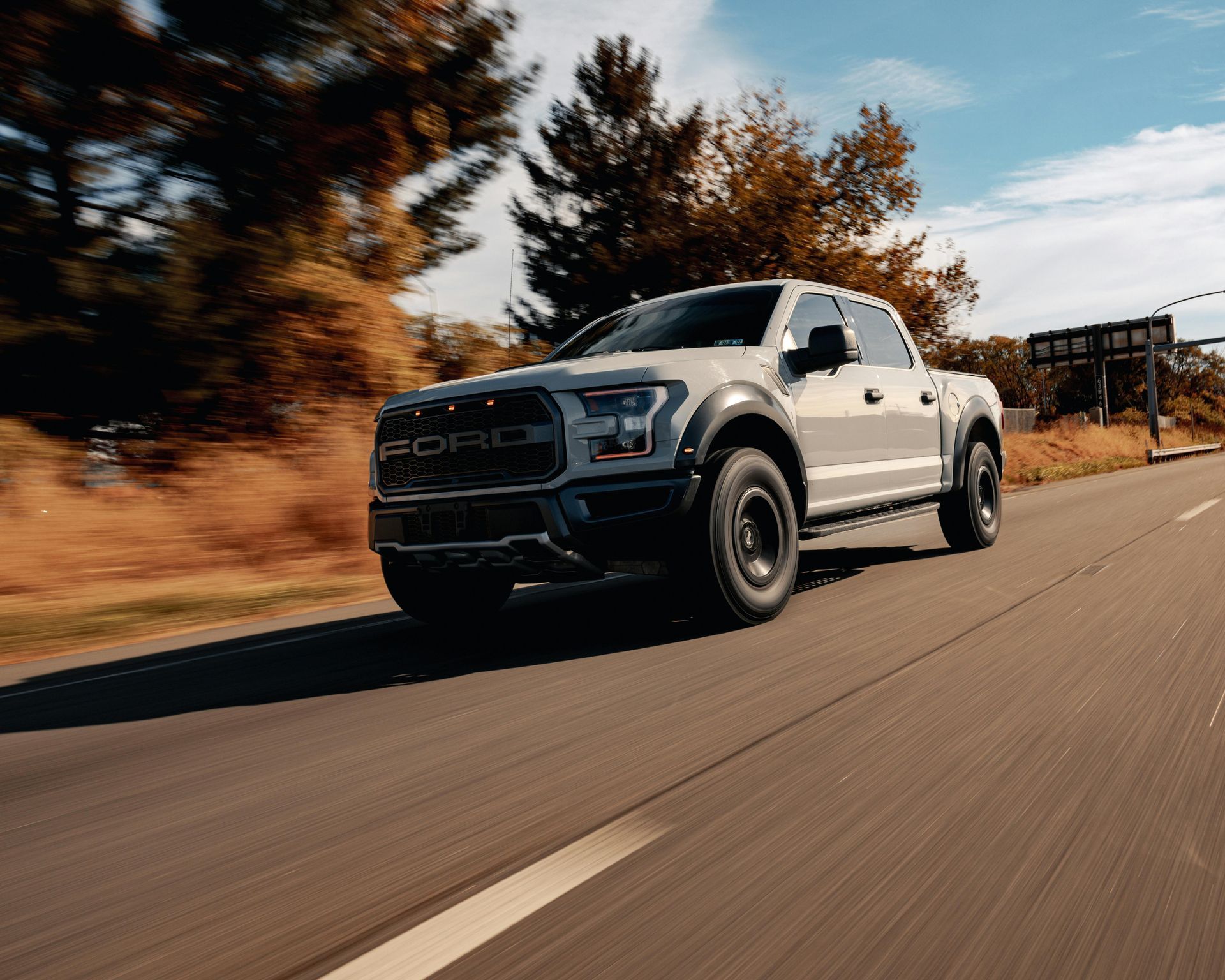 White Ford Raptor truck driving on a road with fall foliage.