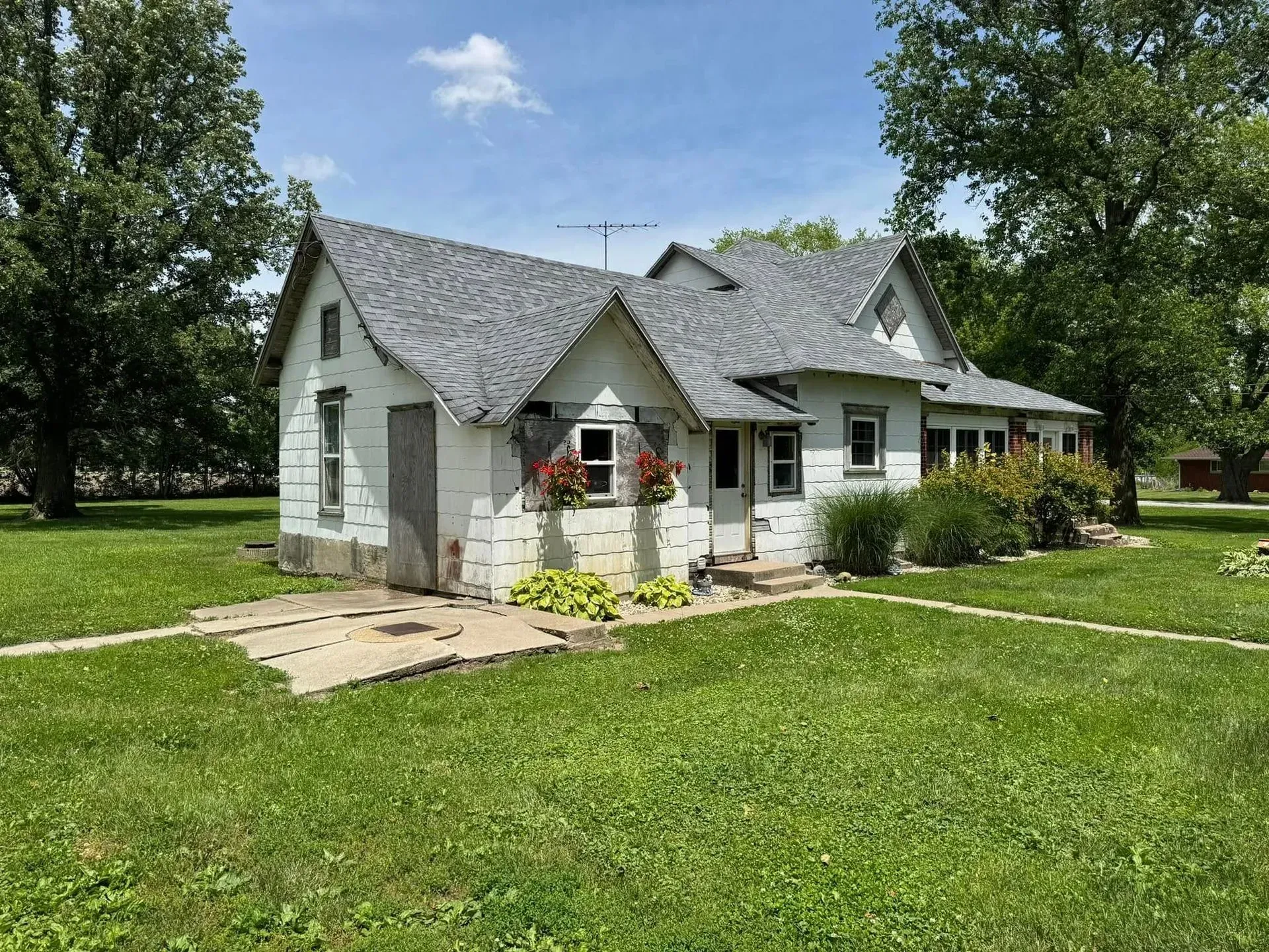 White, weathered house with gray roof, set on green grass under a blue sky with trees.