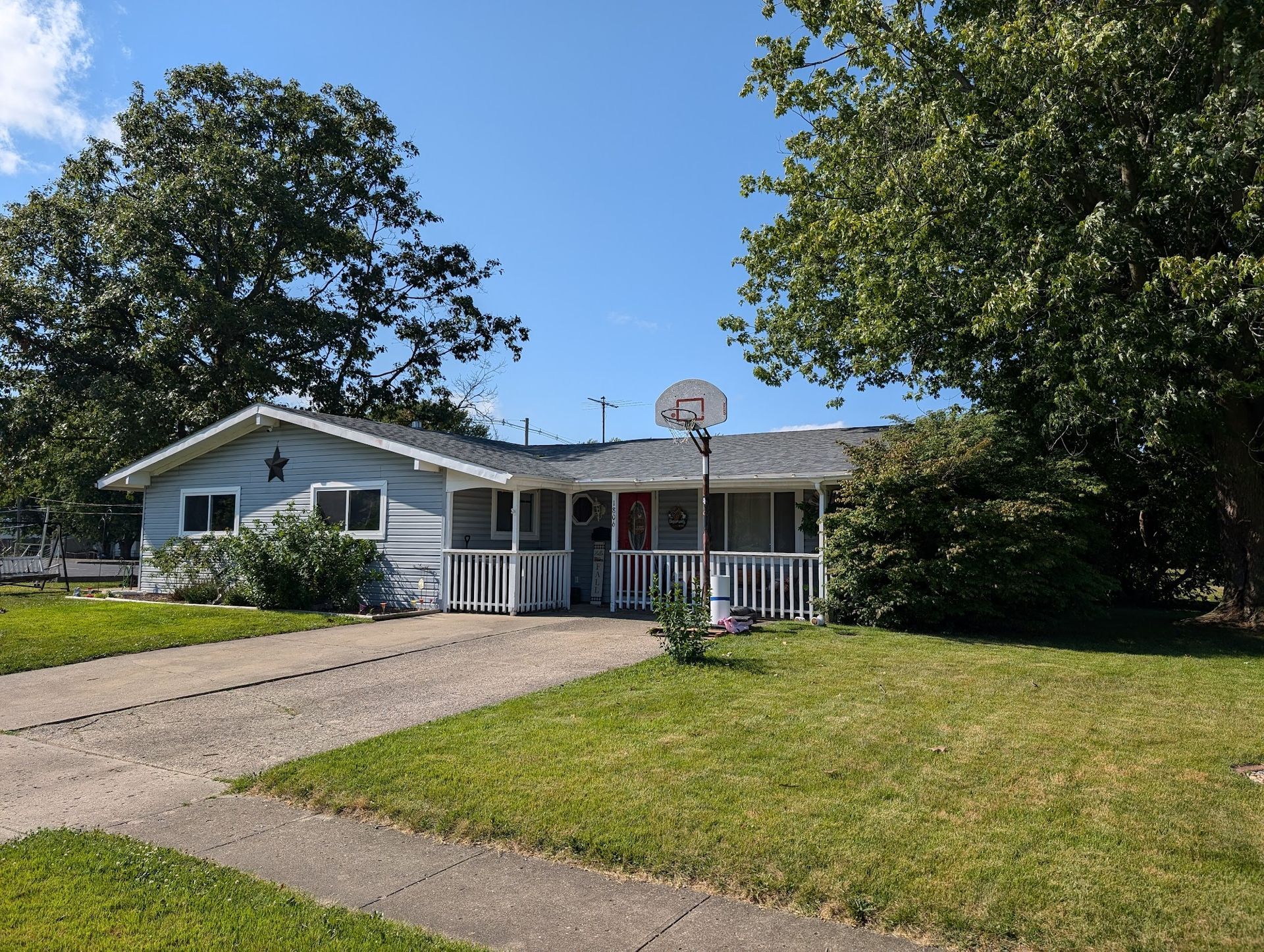 Light blue bungalow house with white porch, driveway, and basketball hoop on a sunny day.