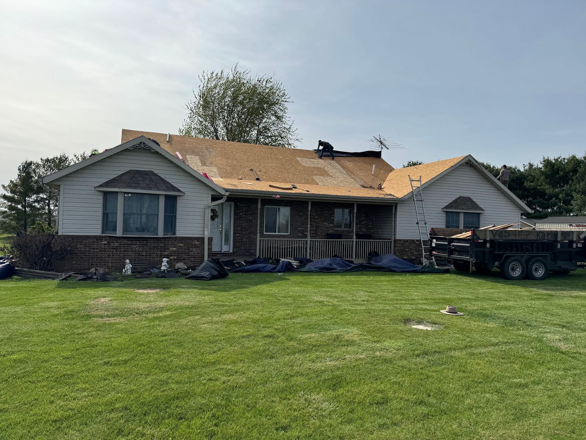 House with roof partially removed, construction underway, surrounded by green grass and a trailer.