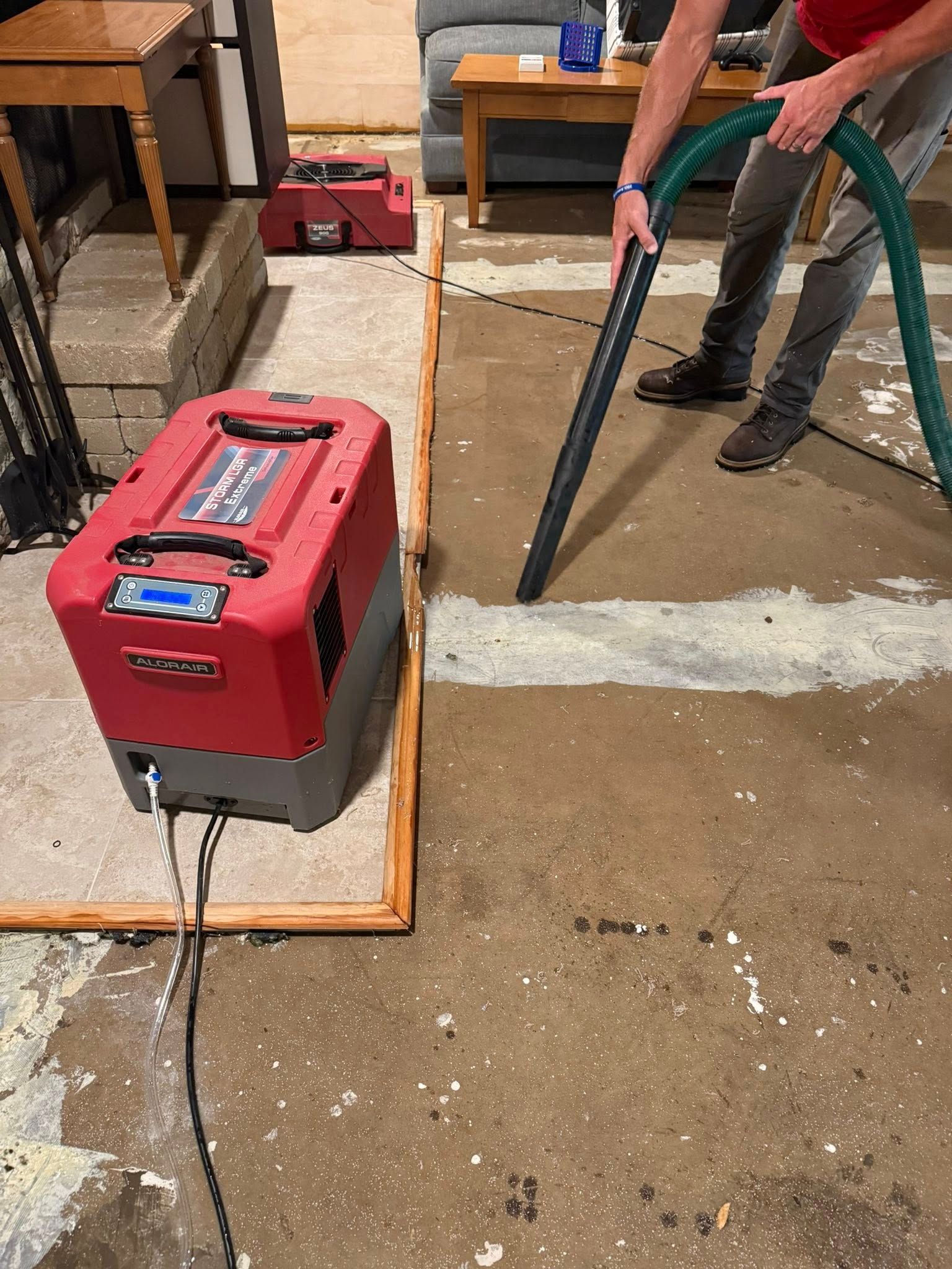 Man using a vacuum hose to clean wet concrete, near red dehumidifiers in a basement.