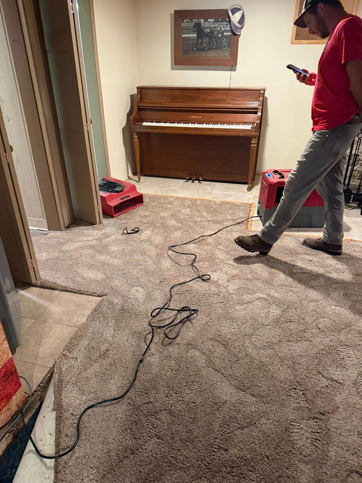 Man in red shirt near a piano, looking at phone. Cord on carpet in room with wooden frame.
