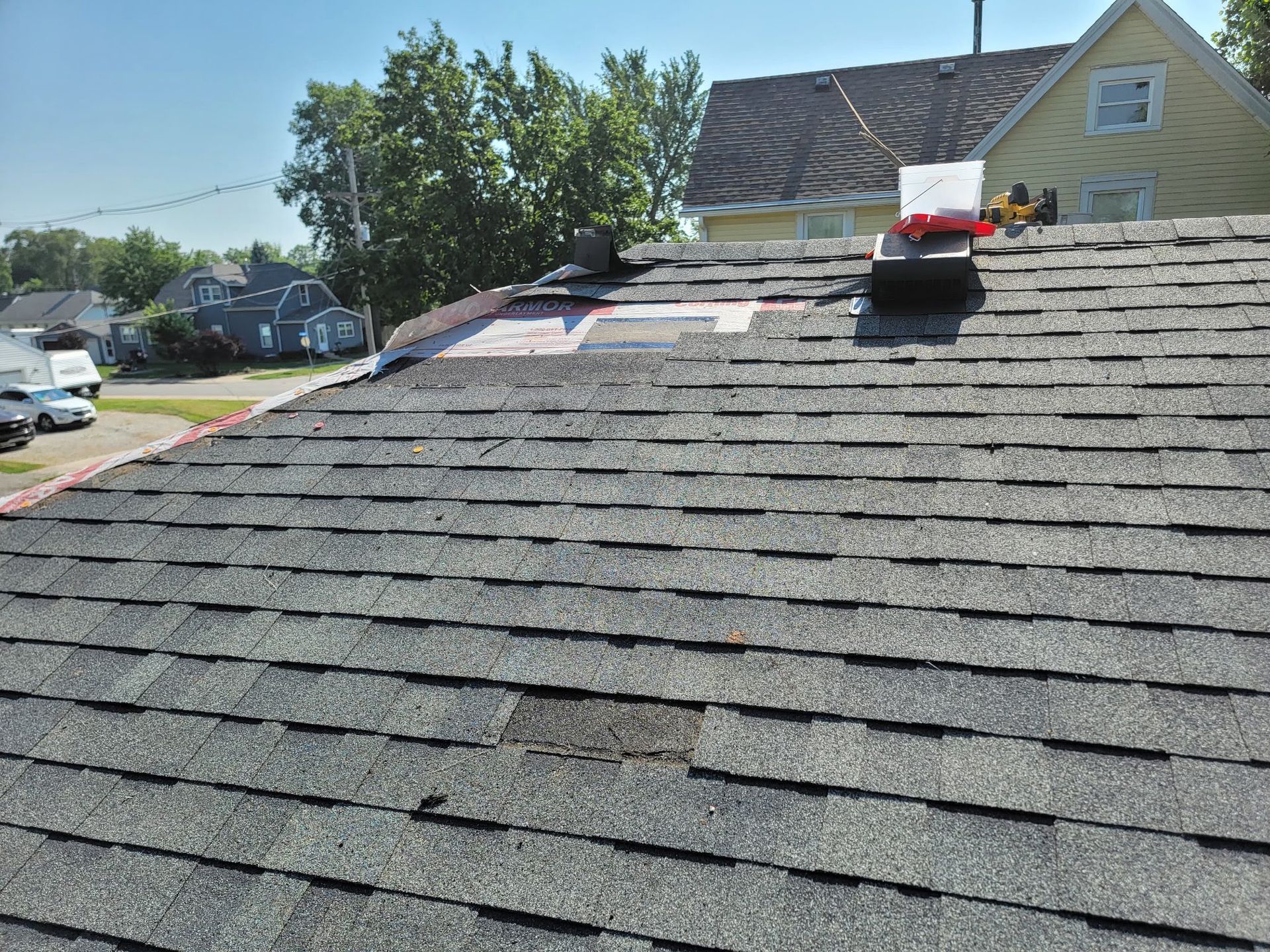 A rooftop with missing shingles, a vent, and a partial repair, in front of houses and trees.
