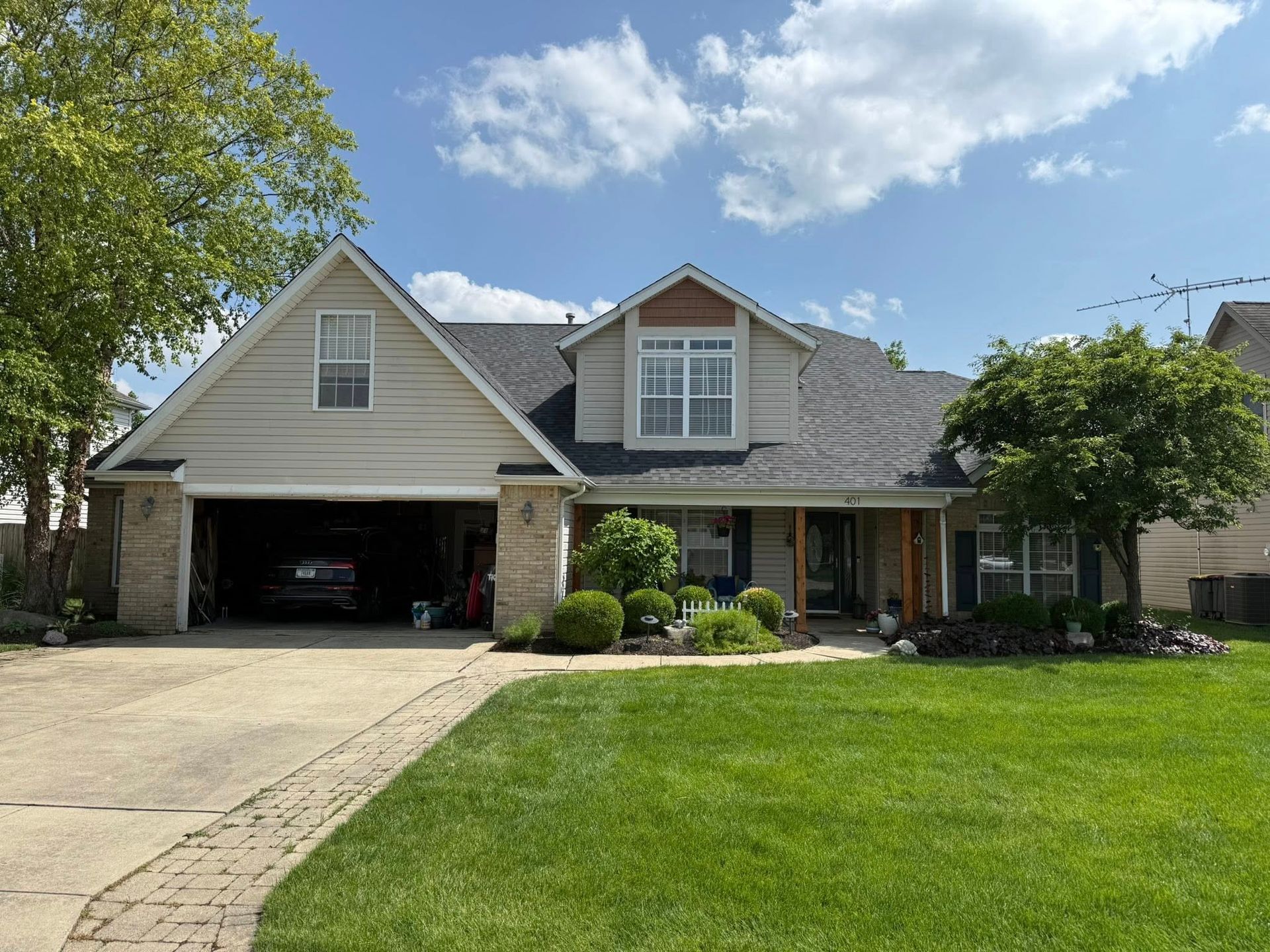 Beige house with a dark roof and a green lawn. The garage door is open, revealing a car. Blue sky.