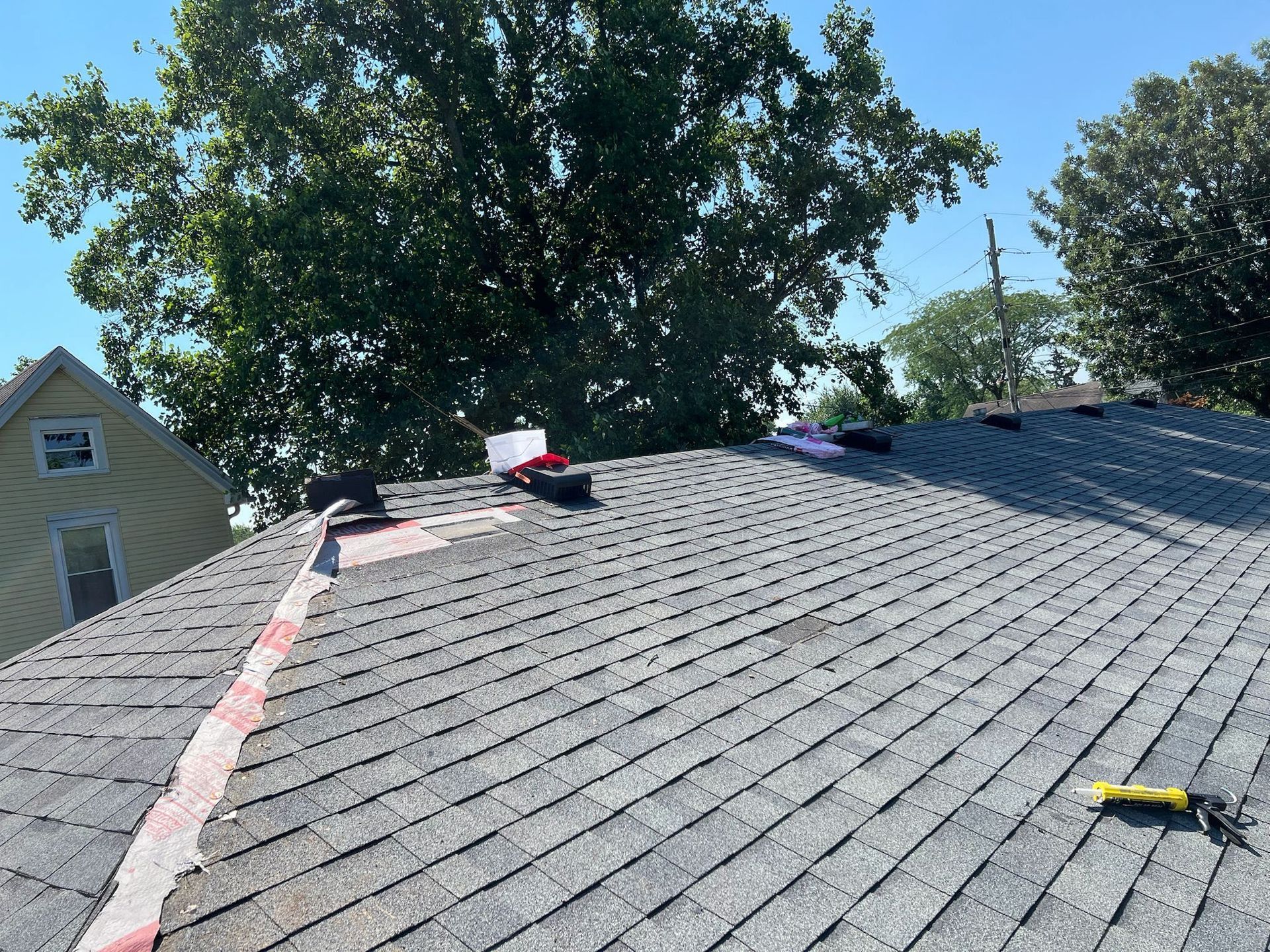 A rooftop with gray shingles and vents, with tools and materials on the surface. Trees and a building are in the background.