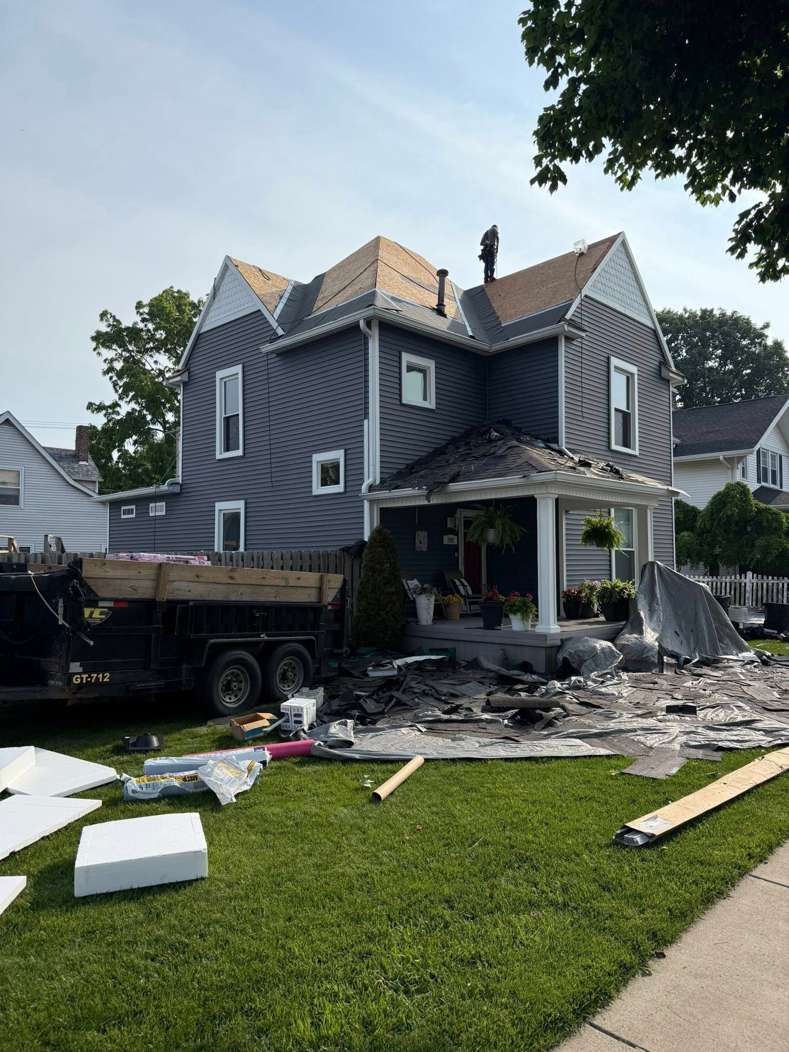 House undergoing roof repair; debris and materials on lawn and in trailer. Gray siding, white trim, sunny day.