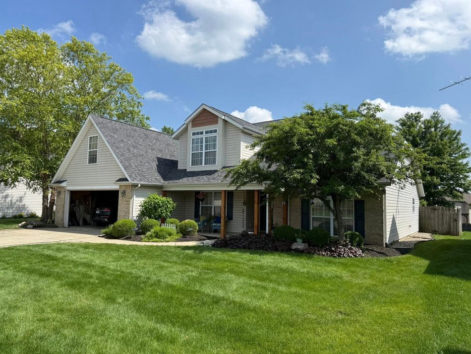 Two-story house with green lawn, trees, and blue sky with clouds. Beige siding, gray roof, and attached garage.