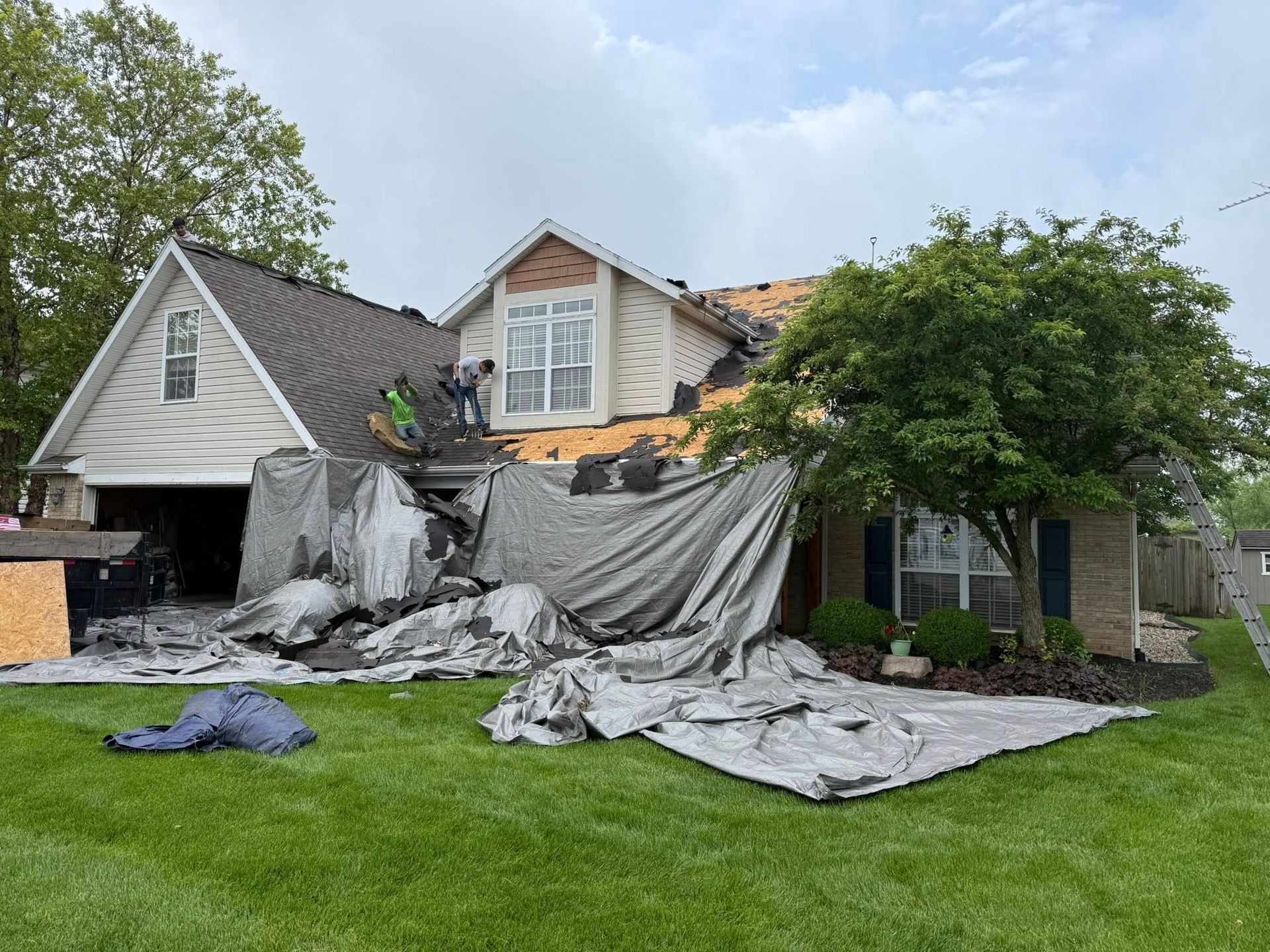 House with roof partially removed, covered by tarps, workers on the roof, green lawn, cloudy sky.
