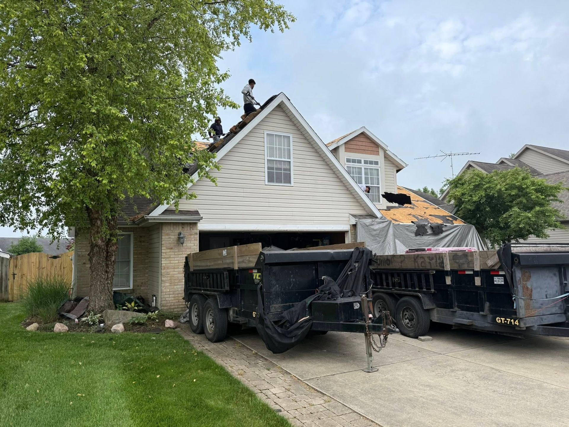 Roofers replacing a roof on a two-story house. Dump trailers parked in the driveway. Green lawn and trees. Cloudy sky.