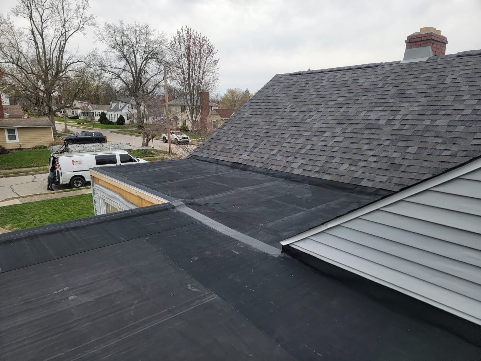 Rooftop view of a home with shingle and flat black roofing. A white van is parked near the house.