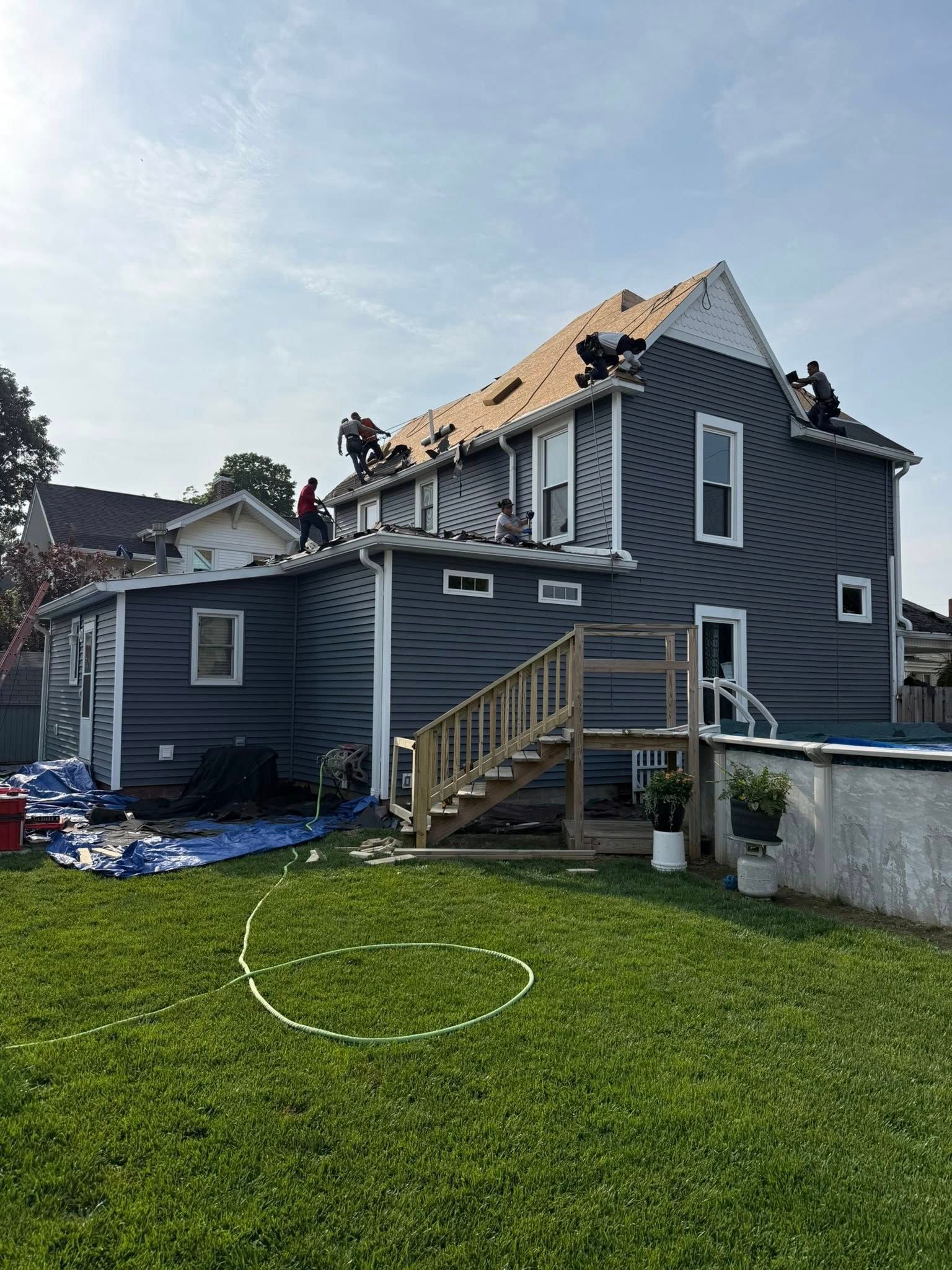 Roofers working on a two-story house with dark gray siding, blue tarps on the ground, and a sunny sky.