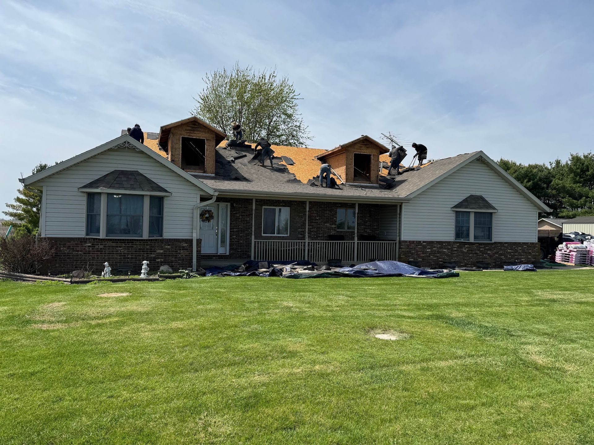 Roofers working on a house with dormers, under a partly cloudy sky.