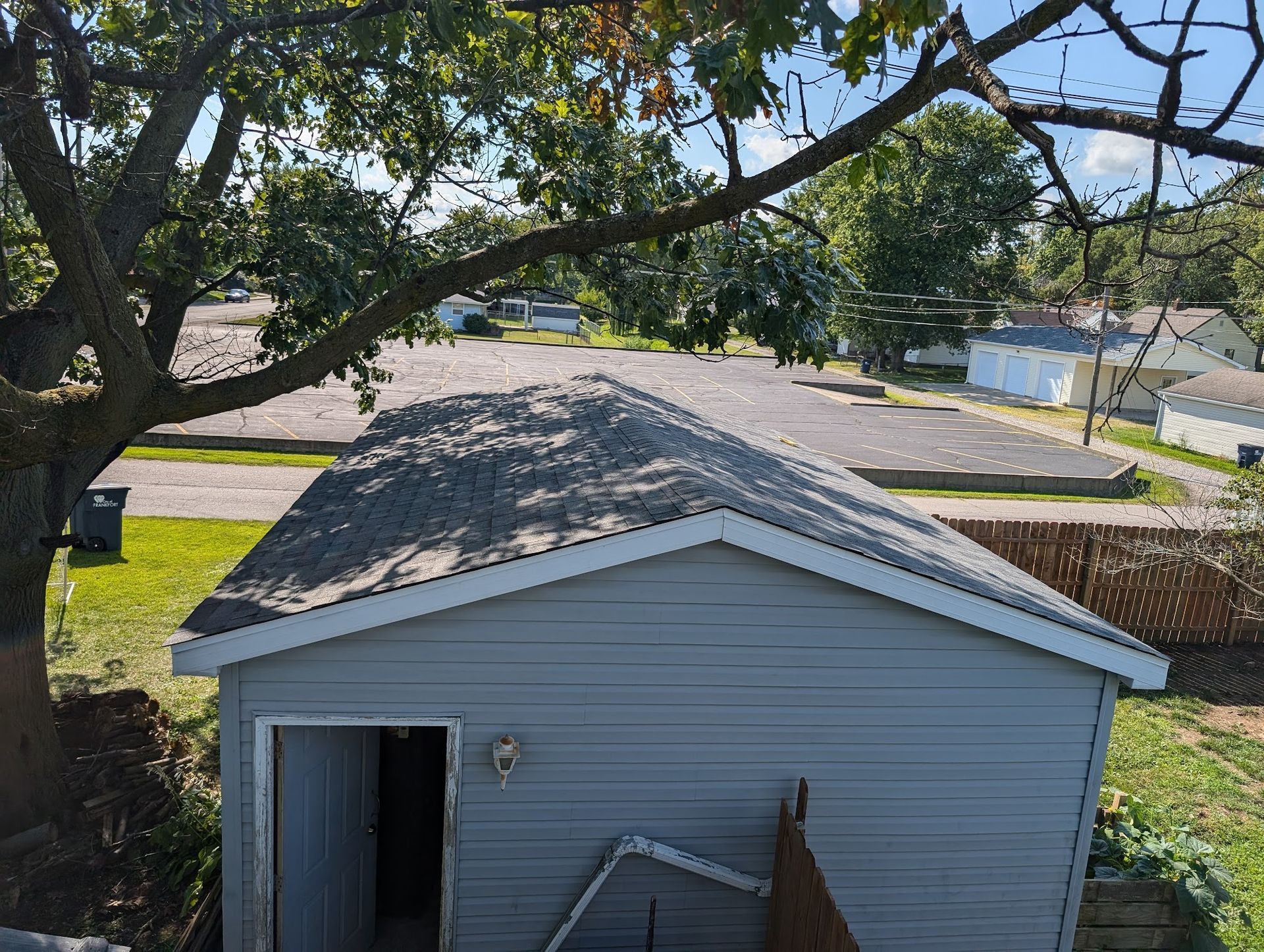 Gray-sided shed with dark roof under tree branches, set in a yard with gravel and other buildings in the background.
