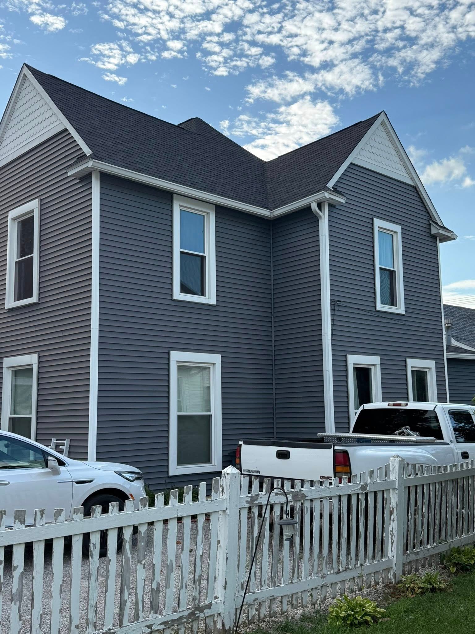 Two-story gray house with white trim and picket fence, parked cars.