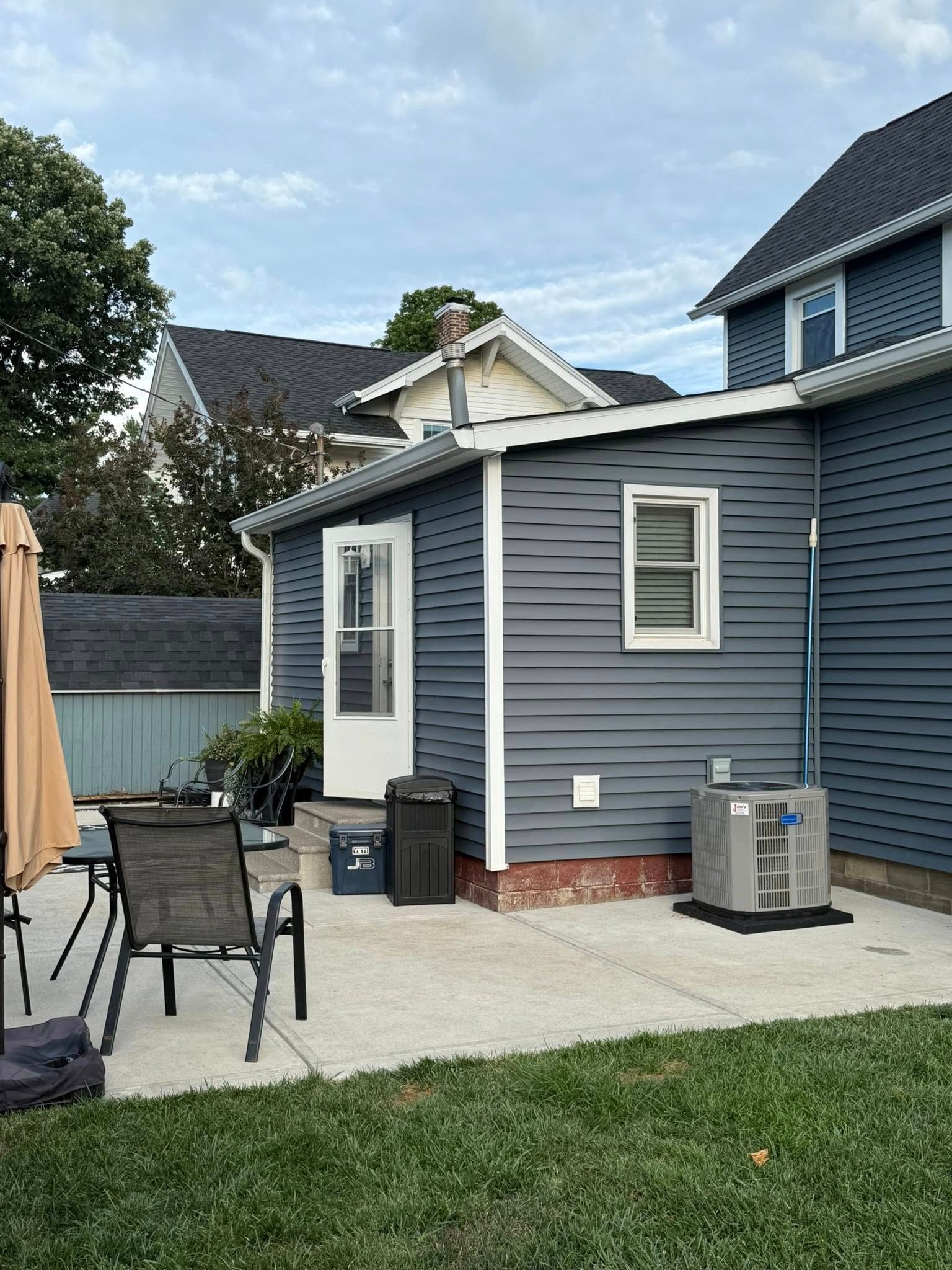 Backyard patio with blue siding, door, window, and air conditioner unit. Green grass and overcast sky.