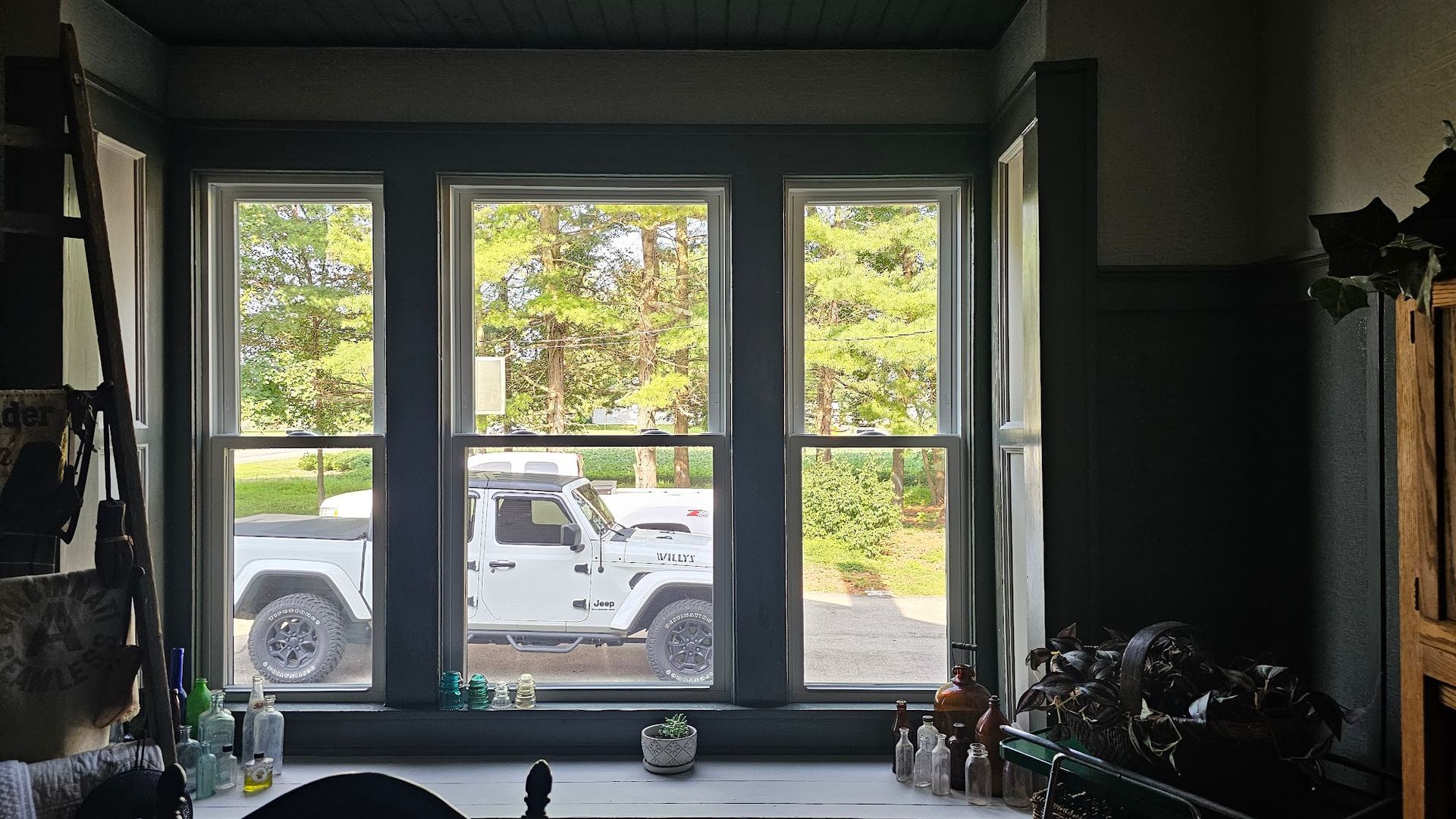 Three-pane window with a white Jeep parked outside, a dark-painted interior, and greenery visible.