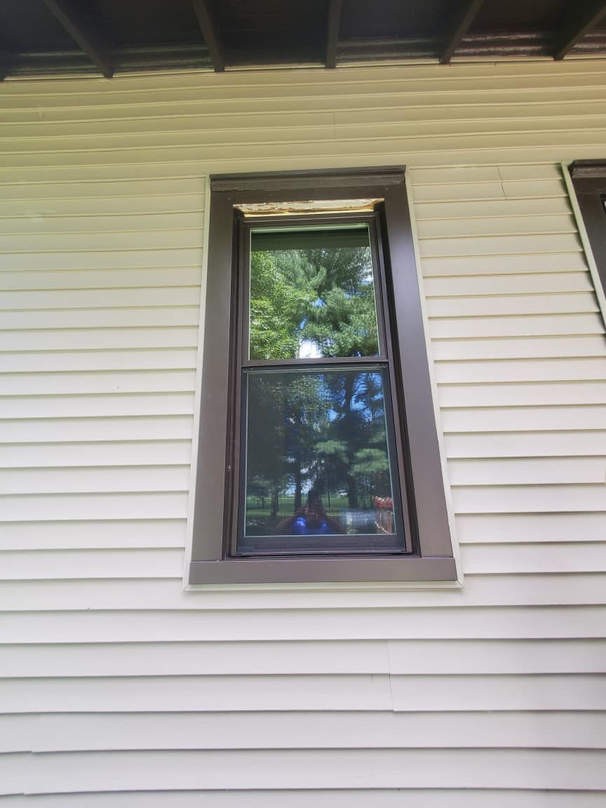 A rectangular window with dark brown trim, reflecting trees and sky, set in a light beige clapboard wall.
