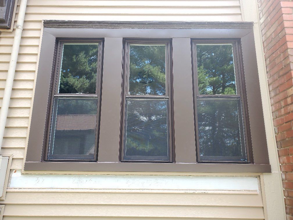 Three brown-framed windows with screens reflecting trees, set in a light brown exterior wall.