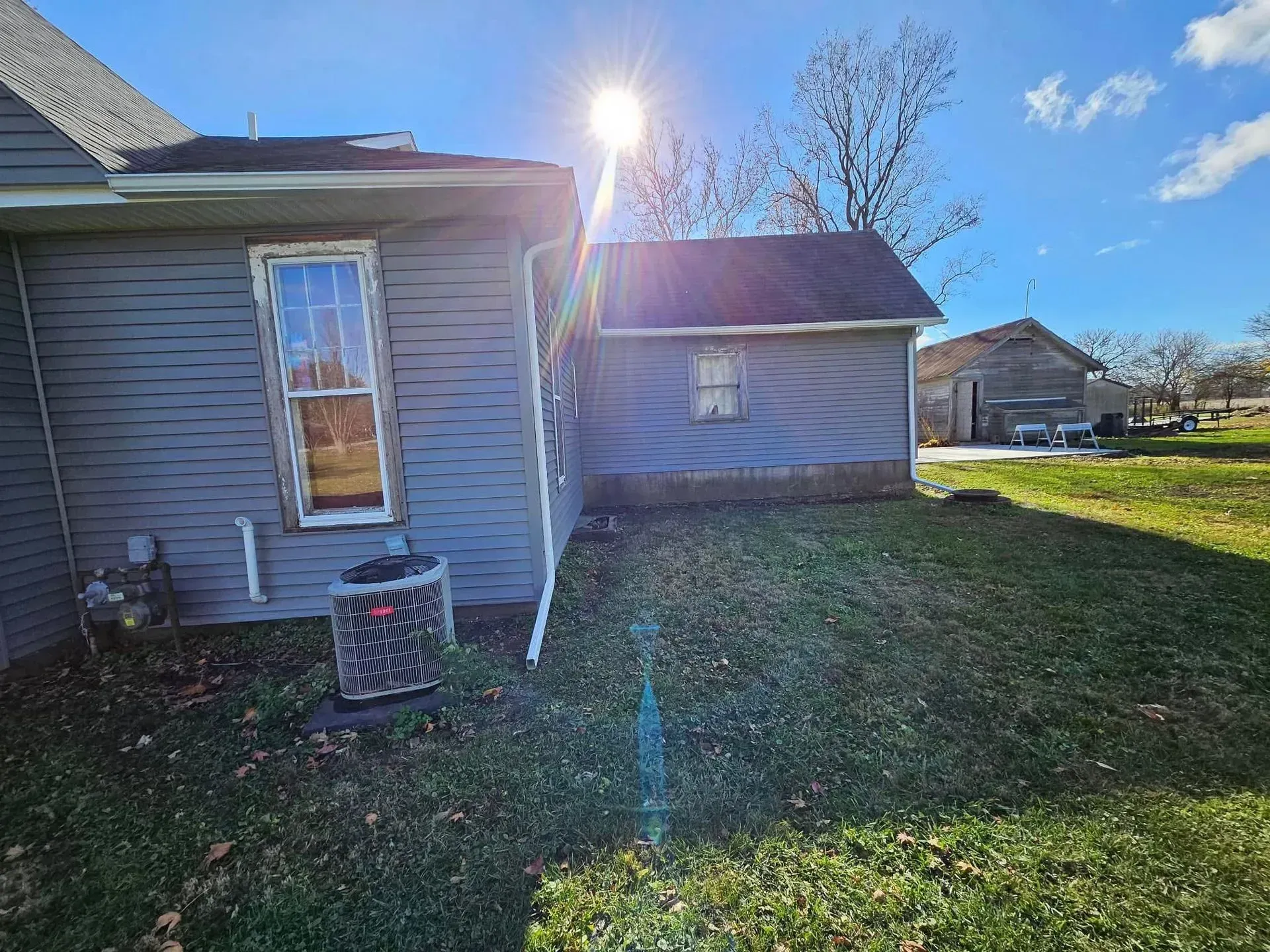 Blue-gray house and shed in a yard on a sunny day. Green grass and trees in the background.